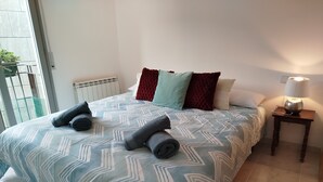 Un dormitorio con una colcha estampada, toallas enrolladas, una lámpara de noche y un radiador.
