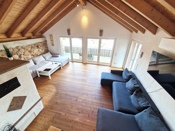 A spacious living area with a high ceiling, wooden beams, a stone wall, a grey sofa, a white coffee table, and a sliding glass door leading to an outdoor balcony.