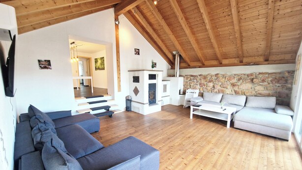 Een ruime woonkamer met houten plafond, stenen muur, open haard en een grijze bank.