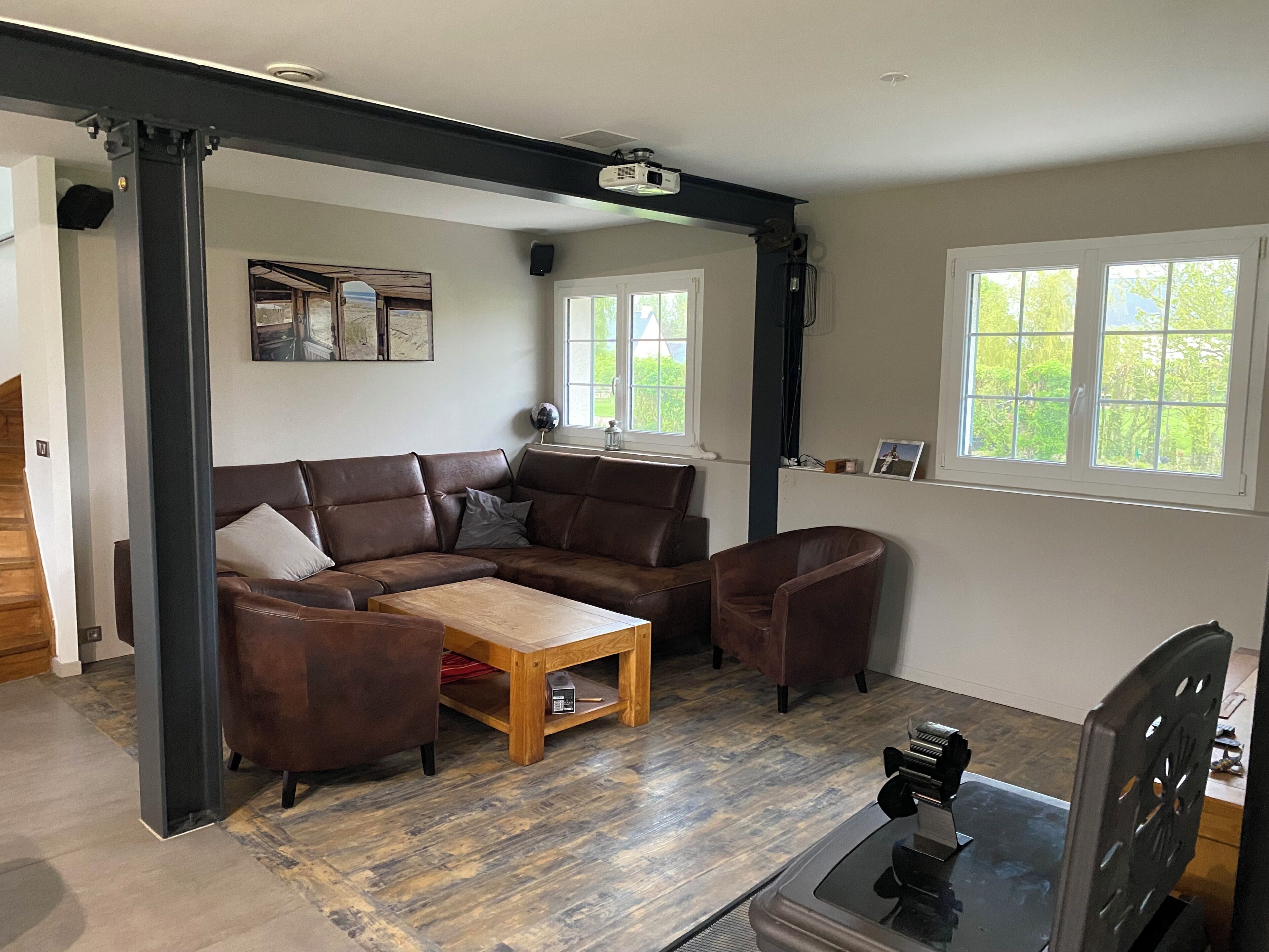 A living room with a sectional sofa, a coffee table, and a television. There are windows with views of greenery outside.