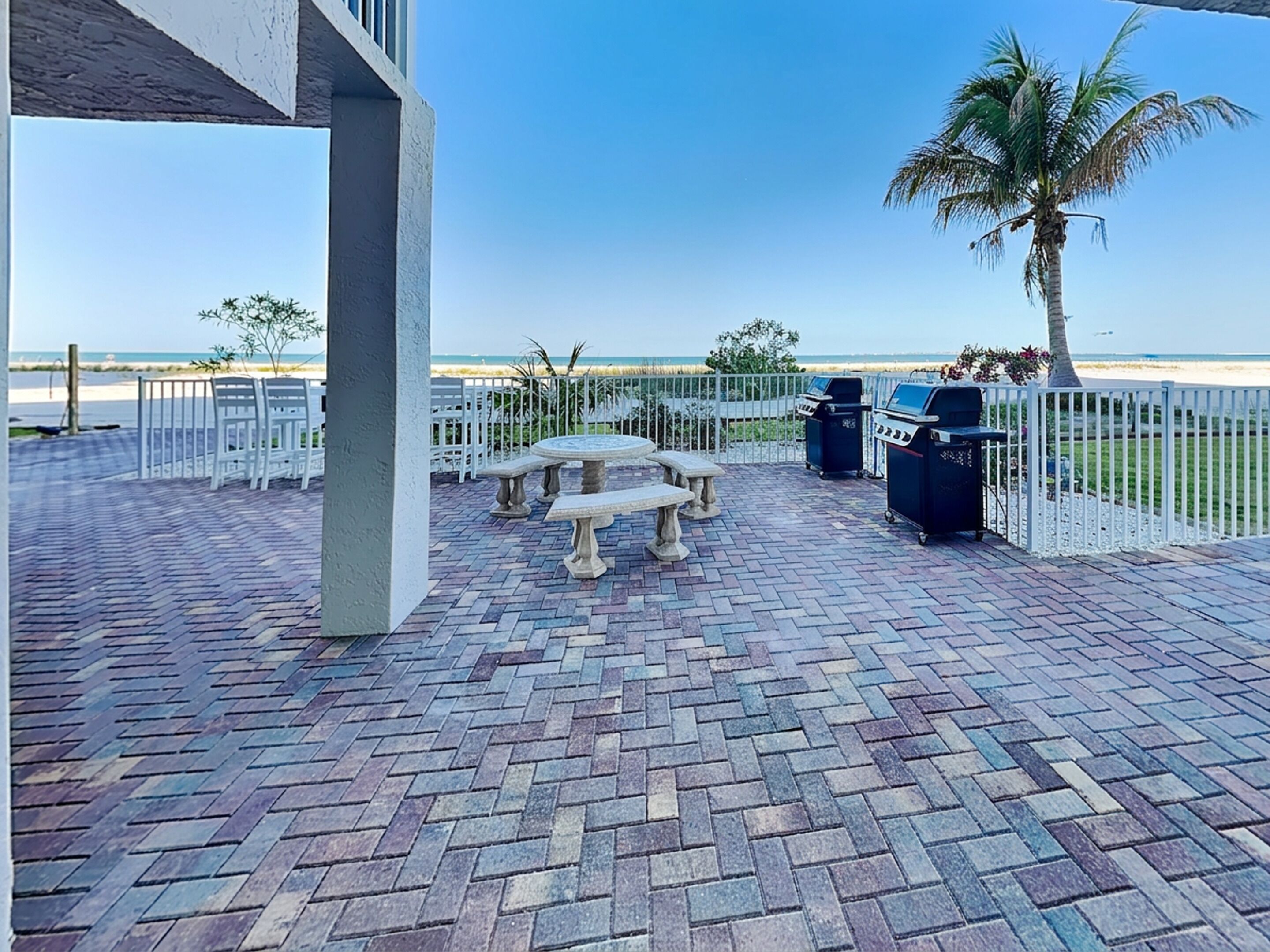 A paved patio with a stone table and benches, a grill, and a palm tree.
