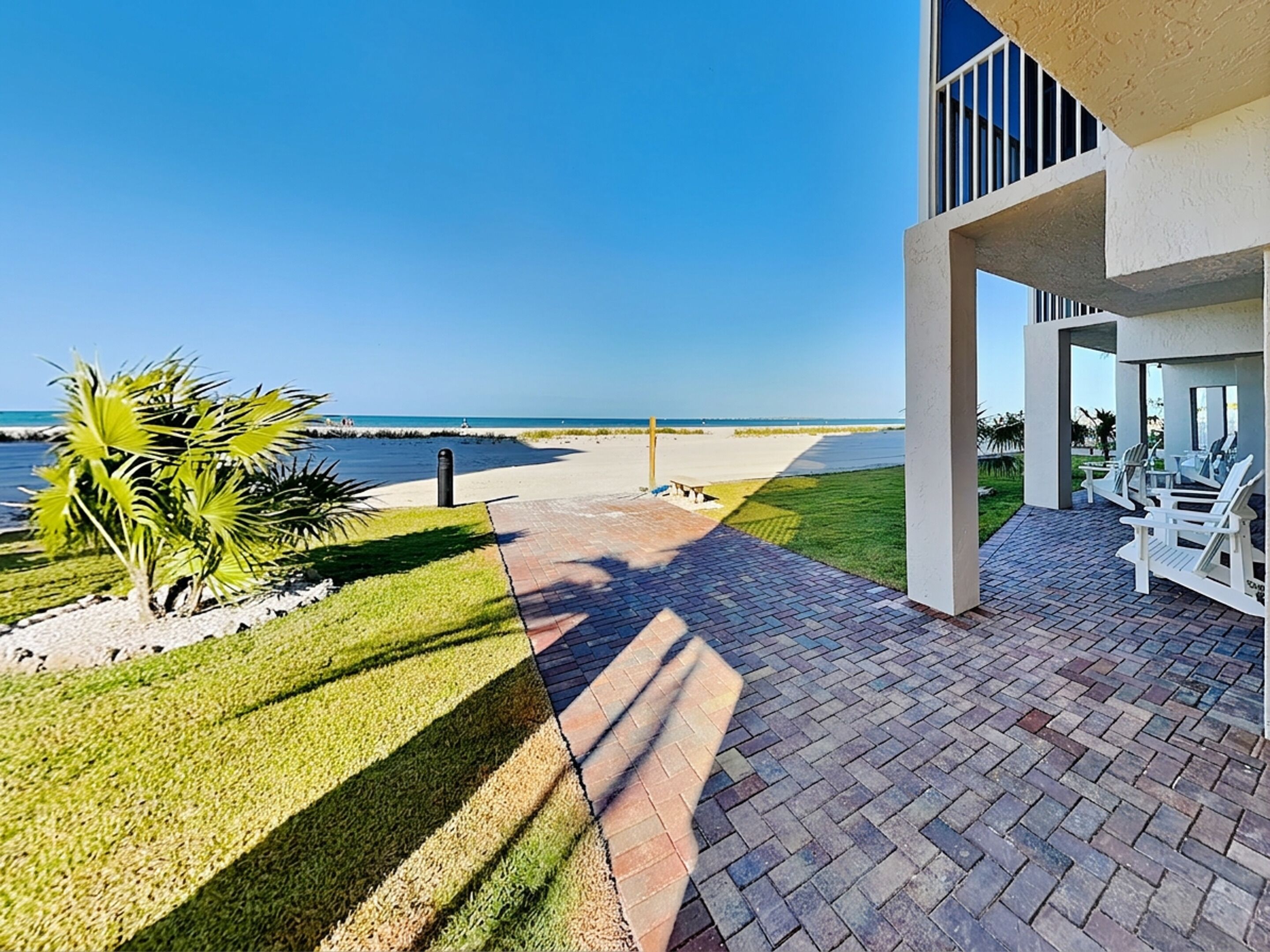 A patio with a view of the ocean, a grassy area with palm trees, and a building with a balcony.