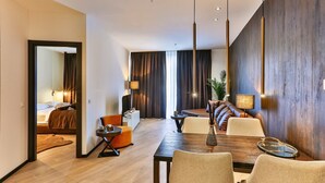 A modern hotel room with a wooden floor, a dining area with a wooden table, and a living area with a sofa and a TV.