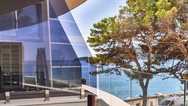 A modern outdoor dining area with a glass table, wine bottle, and glasses, overlooking a body of water and a tree.
