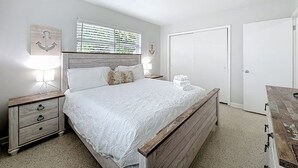 A bedroom with a wooden bed, white bedding, a nightstand with a lamp, a window with blinds, and a door. - Serene 2-bedroom house minutes to Beach Restaurants (Pompano Beach)