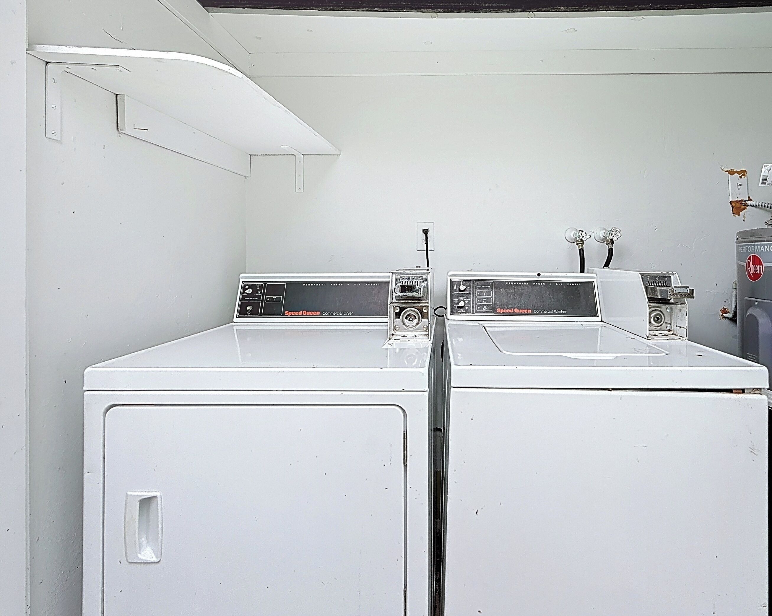 A laundry room with two white washers and dryers, a wall-mounted shelf, and a water heater.