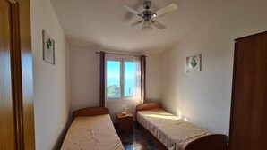 Une chambre avec deux lits simples, un ventilateur de plafond, une fenêtre avec des rideaux et une petite table avec une lampe.