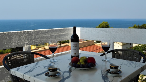 Une table avec une bouteille de vin, des verres et des fruits, placée sur un balcon donnant sur la mer.