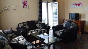 Un salon comprenant un canapé, des fauteuils, une table basse en verre, un téléviseur et une porte-fenêtre coulissante donnant sur un espace extérieur.
