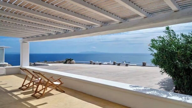 Een overdekt terras met uitzicht op zee, houten meubels en een witte balustrade.