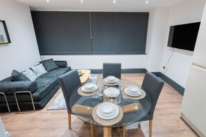 Un salon moderne avec une table à manger en verre pour quatre personnes, un canapé de couleur sombre et un téléviseur à écran plat fixé au mur.