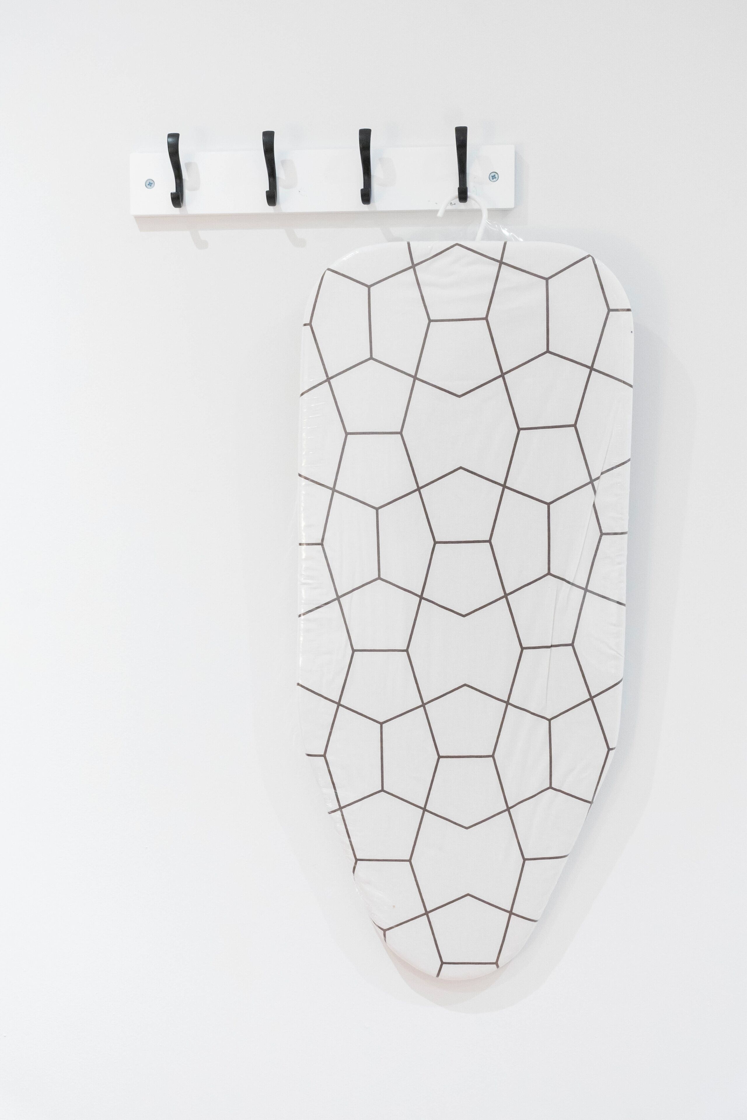 Una tabla de planchar blanca con un diseño geométrico, fijada a la pared con tres ganchos negros.
