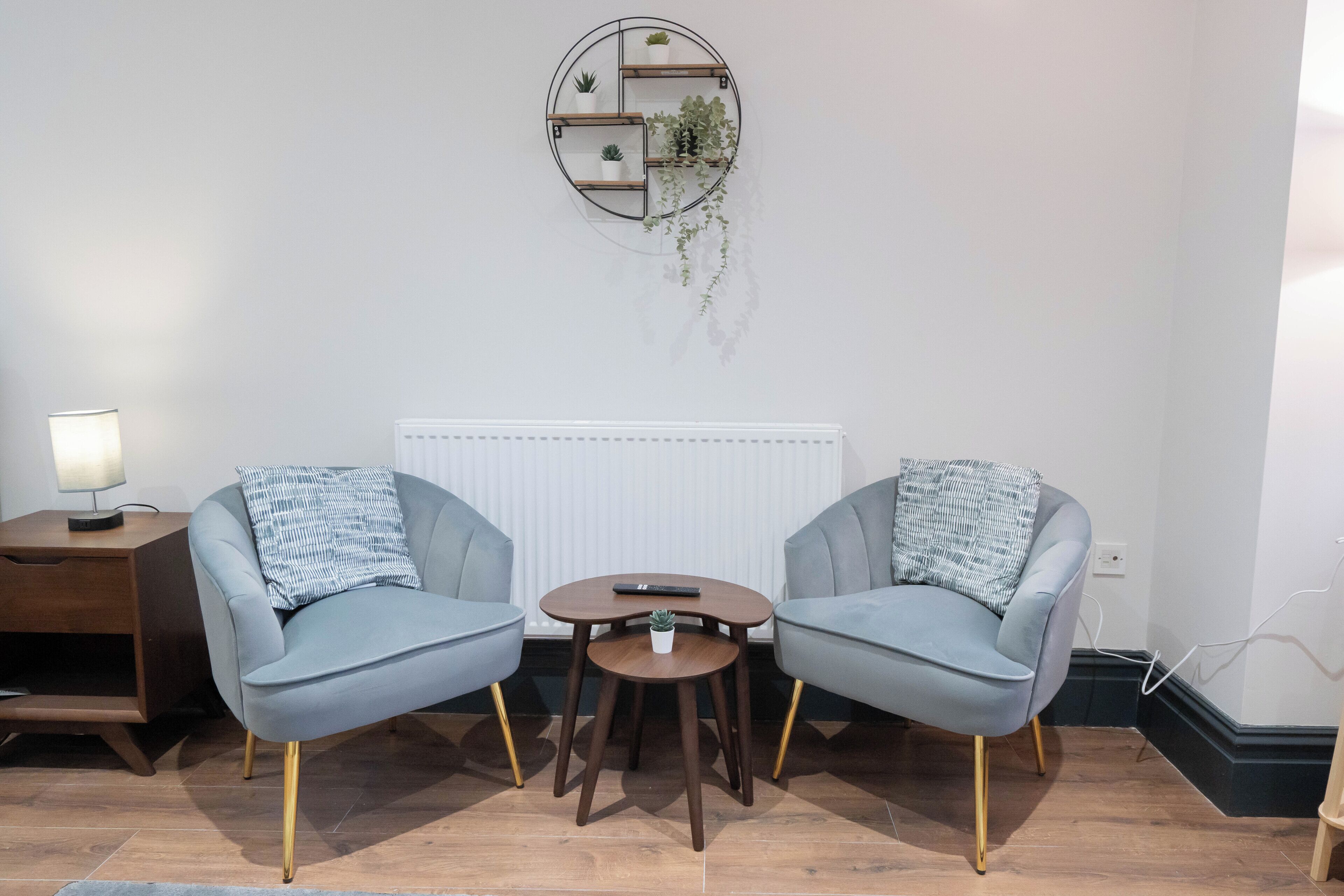 Une pièce avec deux fauteuils gris, une petite table ronde, une étagère murale ornée de plantes, une lampe et un radiateur.