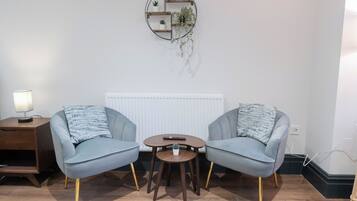 Une pièce avec deux fauteuils gris, une petite table ronde, une étagère murale ornée de plantes, une lampe et un radiateur.