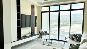 Een moderne woonkamer met een flatscreen-televisie, een eettafel met stoelen, een bank en grote ramen met uitzicht op de stad.