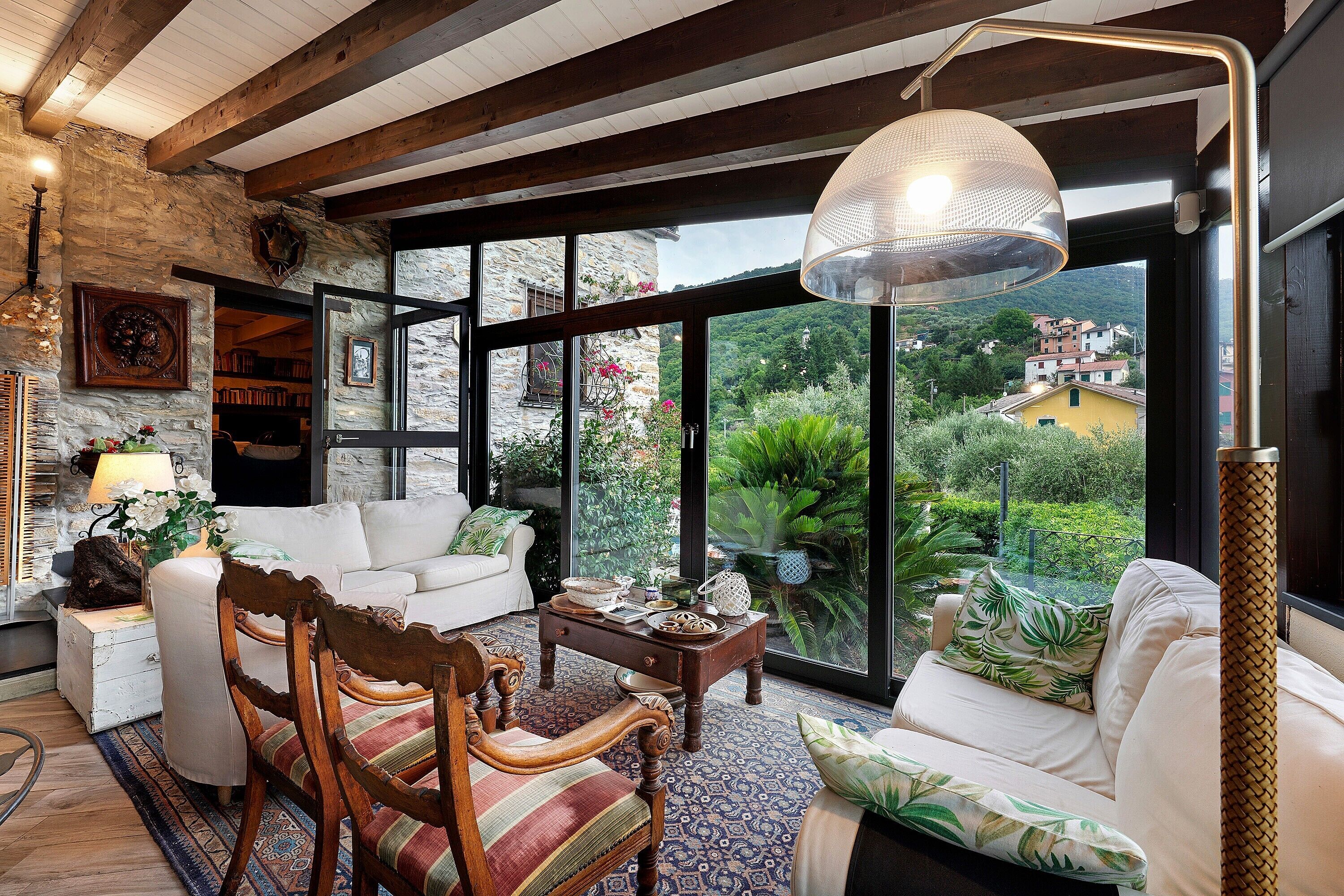 Ein gemütliches Wohnzimmer mit Steinwand, Holzbalken, einem weißen Sofa, einem gemusterten Teppich und einem großen Fenster mit Blick auf Grünflächen und Gebäude.