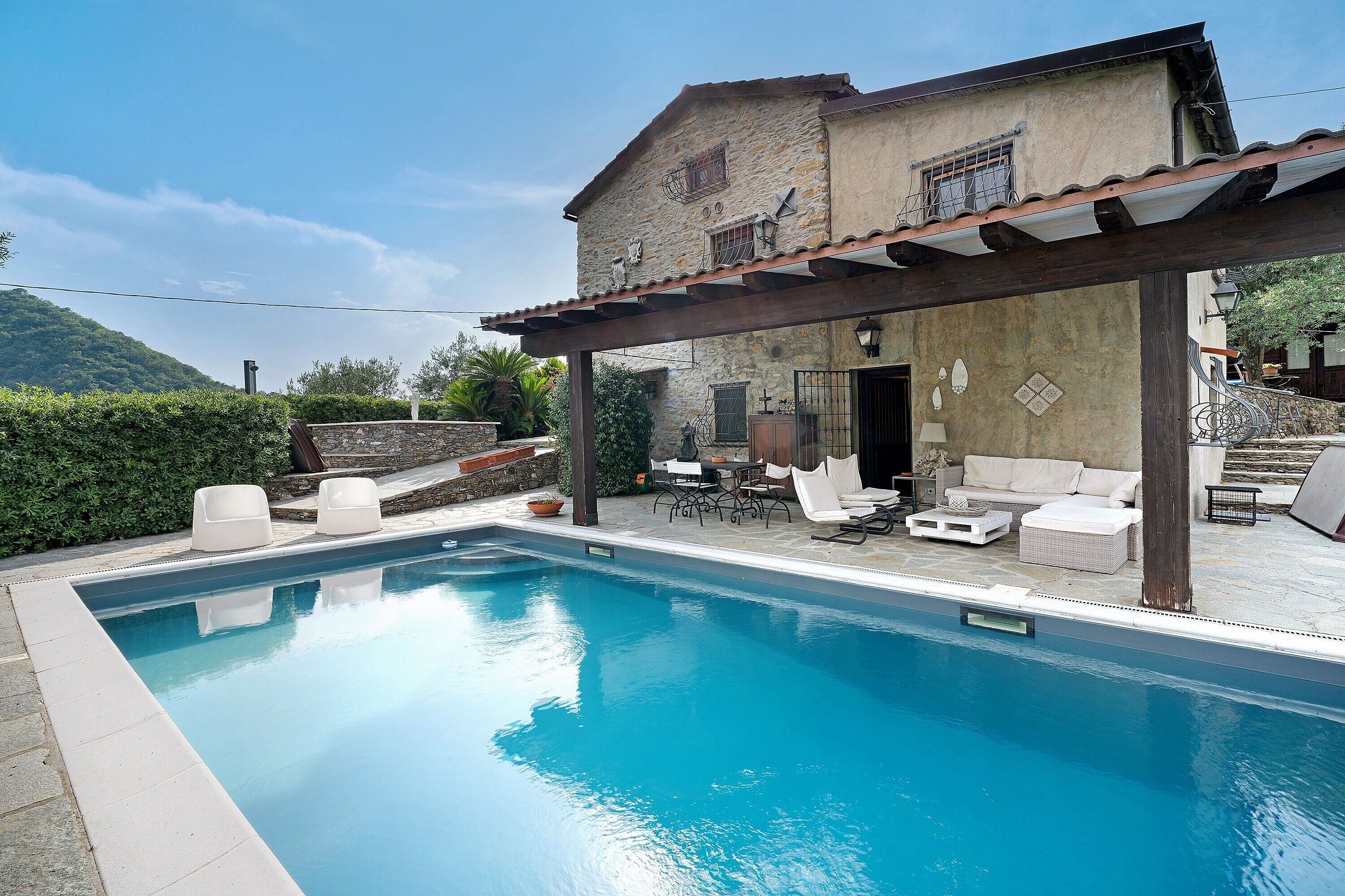 A pool with a pergola and outdoor furniture, surrounded by a stone building with a rustic appearance.