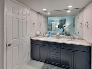 A bathroom with a double sink, dark cabinets, a large mirror, and a visible bedroom in the background.