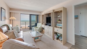 A spacious living room with a large TV, comfortable seating, a glass coffee table, and a view of the ocean through a sliding glass door. - Indies House 307 (Ormond Beach)