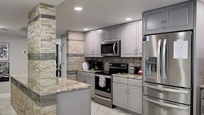 A modern kitchen with granite countertops, stainless steel appliances, and a stone accent wall. - Indies House 307 (Ormond Beach)