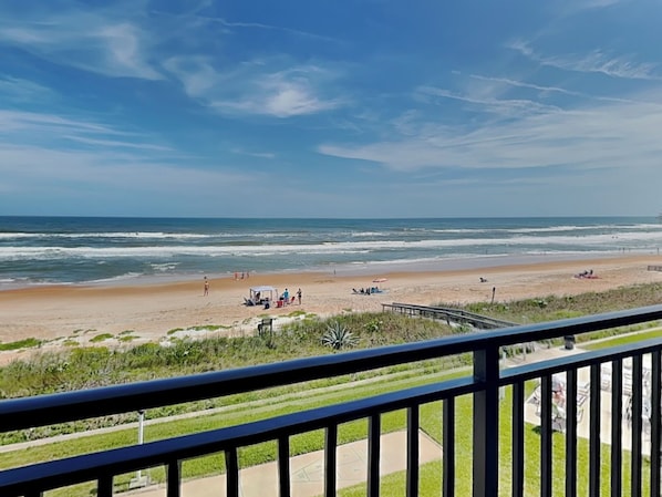 A beach view from a balcony with a railing, ocean, and people on the sand.