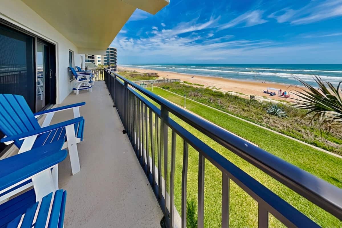 A balcony with blue chairs, a view of the beach, and a clear sky.