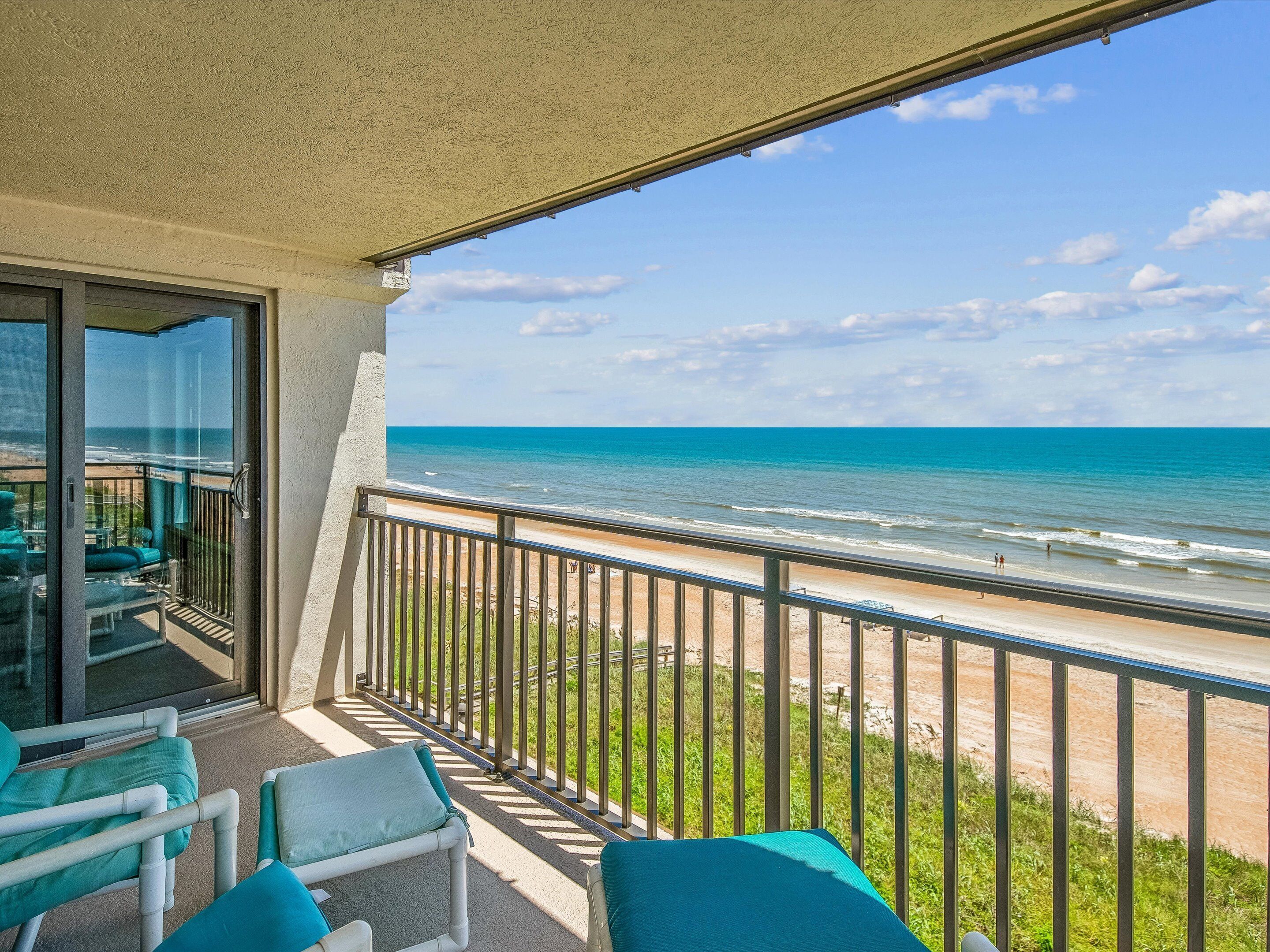 A balcony with a view of the beach, a glass door, and two chairs with cushions.