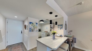 A modern kitchen with a central island, pendant lighting, and a view into the dining area. - Indies House 307 (Ormond Beach)