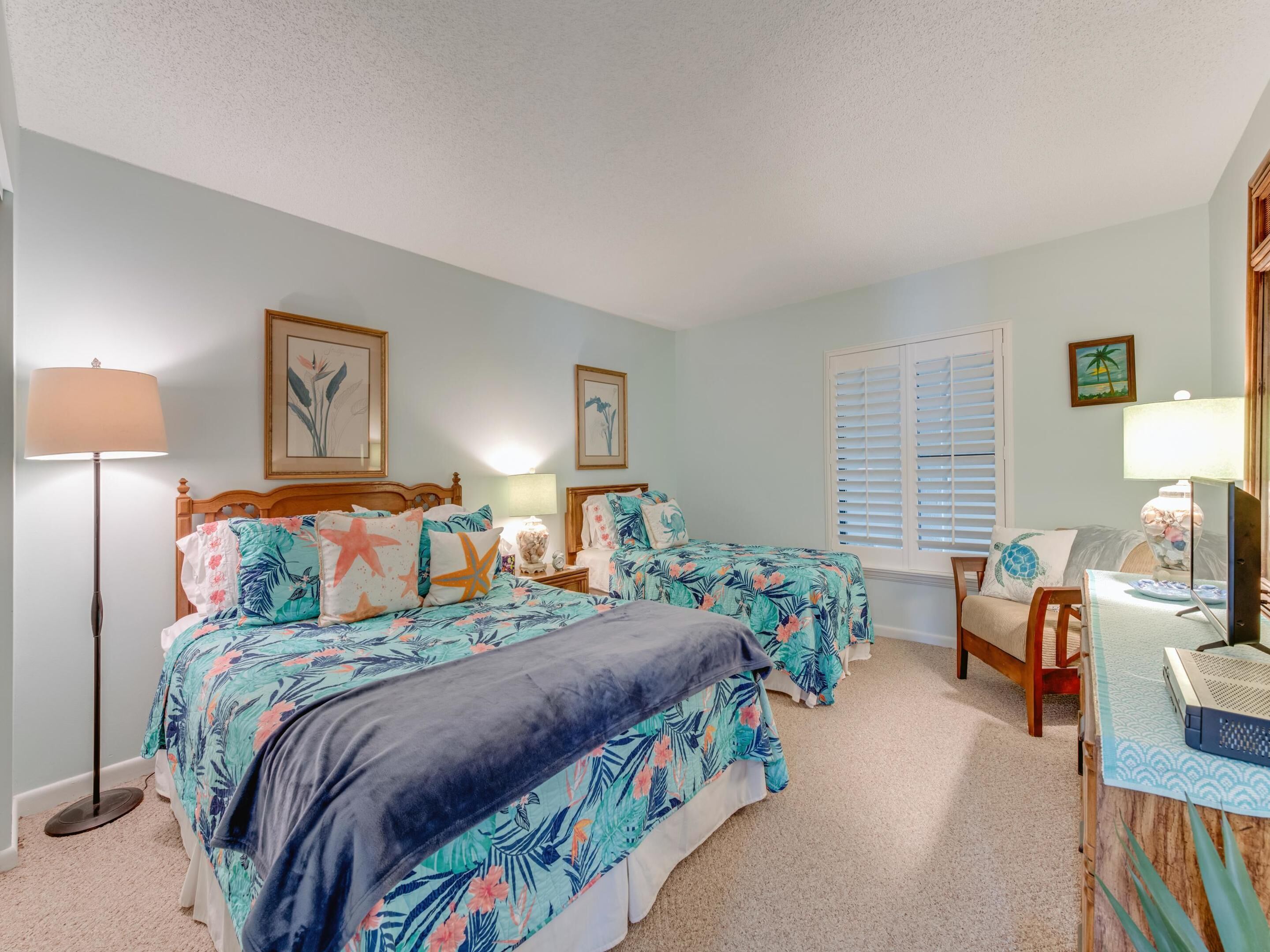 A bedroom with two beds, a desk, a chair, a lamp, and a window with shutters.