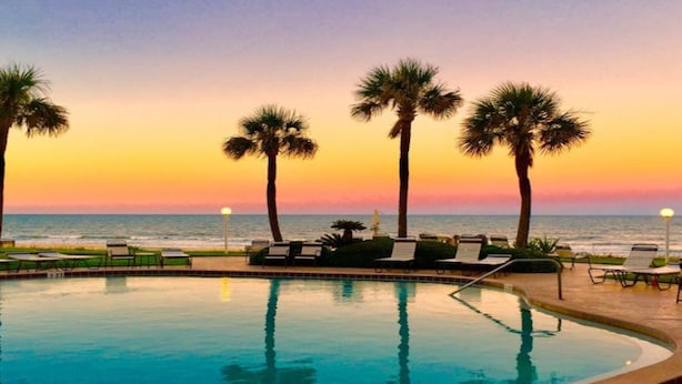 A pool with lounge chairs and palm trees, overlooking the ocean at sunset. - Indies House 307 (Ormond Beach)