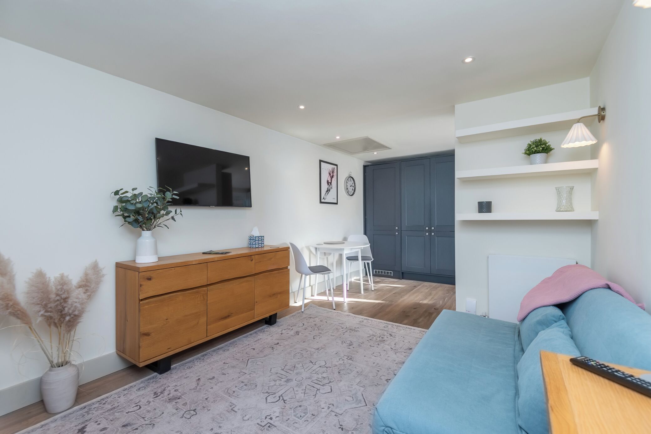 A modern living room with a wooden cabinet, a flat-screen TV, a dining area with a white table and chairs, and a blue sofa.