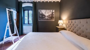 Ein Schlafzimmer mit Bett, einem gerahmten Bild an der Wand, einem Nachttisch mit Lampe und einem Fenster mit Vorhängen.
