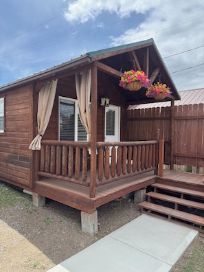 A wooden cabin with a porch, a concrete pathway, and hanging flower baskets. - Tiny Yellowstone Riverside Cabin (Gardiner)