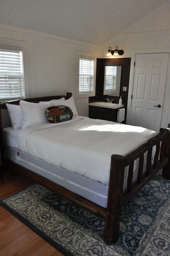 A bedroom with a wooden bed, white bedding, a bedside table, a mirror, and a window with blinds.