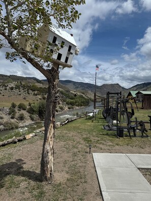 A tree with a birdhouse, a river, a workout area with equipment, and a flag. - Tiny Yellowstone Riverside Cabin (Gardiner)
