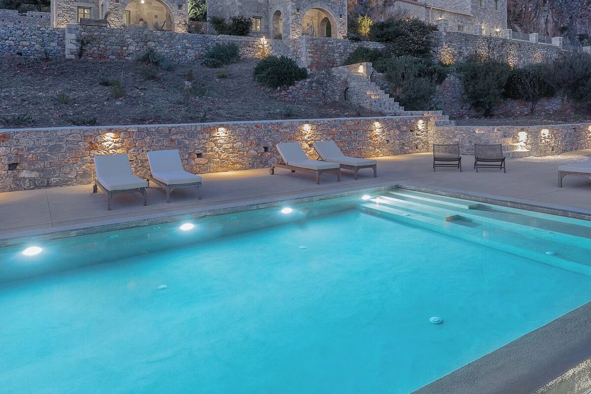 Ein Pool mit Liegestühlen, Steingebäude und eine Bergkulisse im Hintergrund.