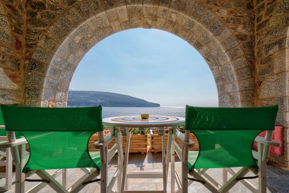 Ein Tisch mit zwei grünen Stühlen vor einem steinernen Torbogen mit Blick auf ein Gewässer.