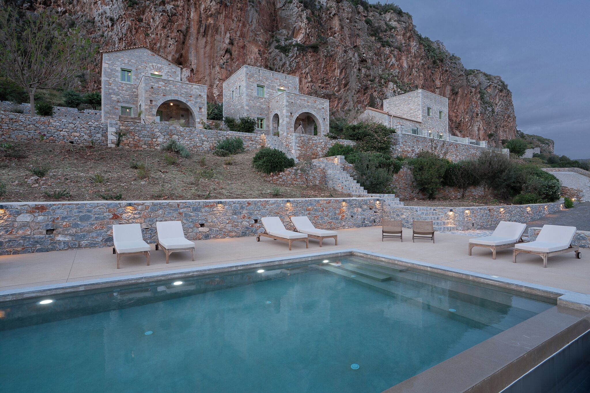 A pool area with lounge chairs, a stone building with arches, and a rocky cliff backdrop.