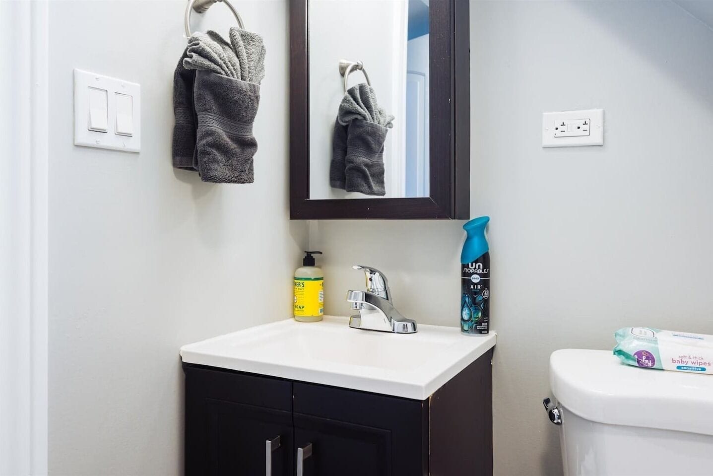 Un bagno con un lavandino bianco, uno specchio, un portasciuga, una bottiglia di sapone, un flacone spray e una confezione di salviette per bambini.