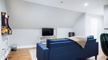A modern living room with a blue sofa, a flat-screen TV on a white stand, a gaming console, and a trash bin.
