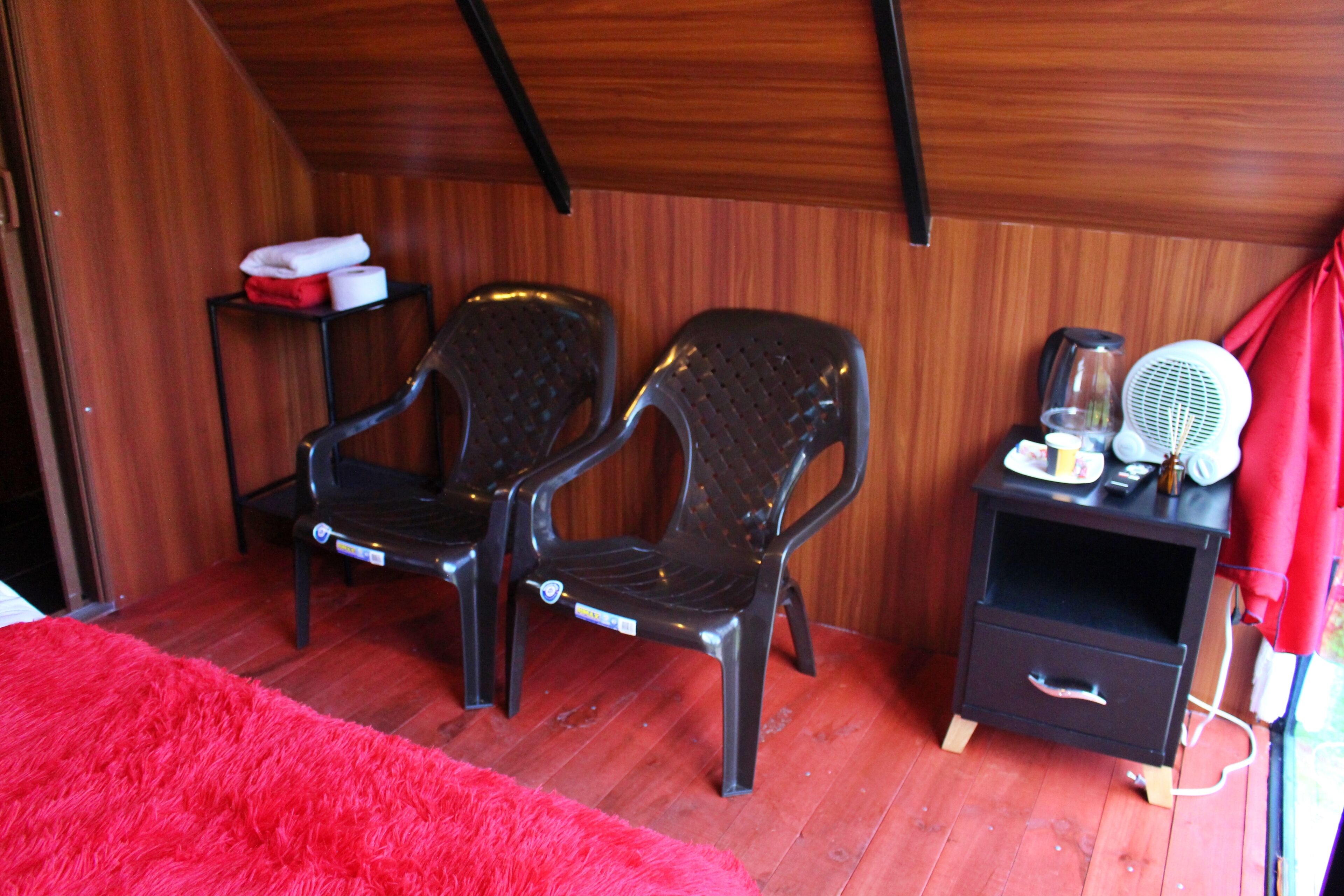 Una habitación con dos sillas de plástico negro, una mesita de noche pequeña con un ventilador, una alfombra roja y paredes de madera.