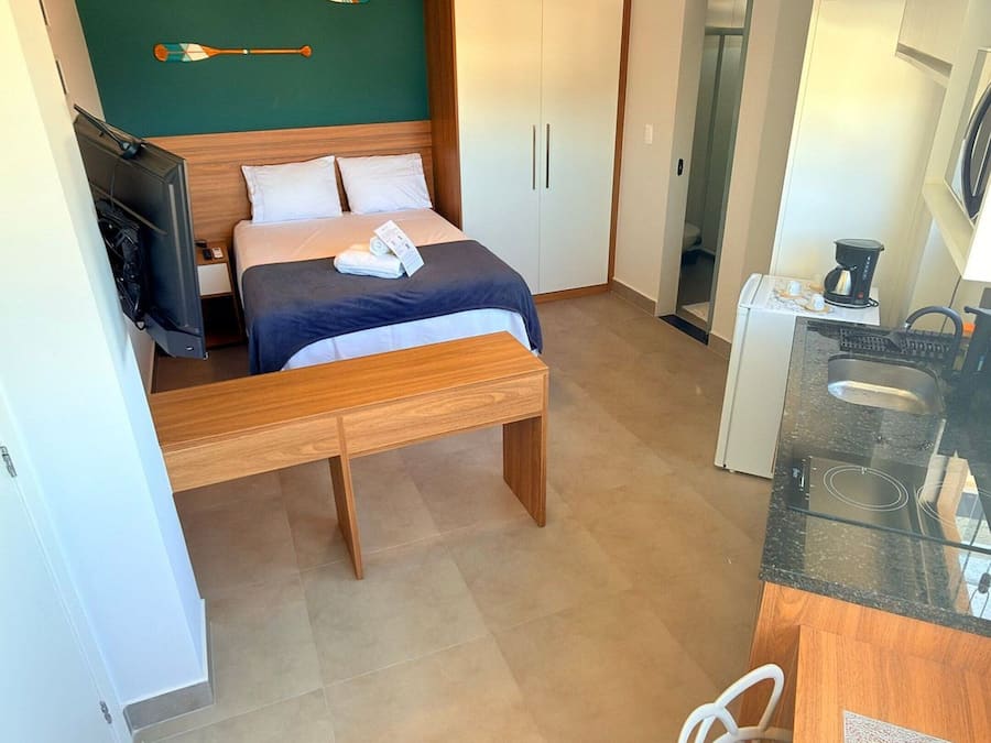 A compact hotel room with a bed, a small desk, a chair, a kitchenette, and a wardrobe.