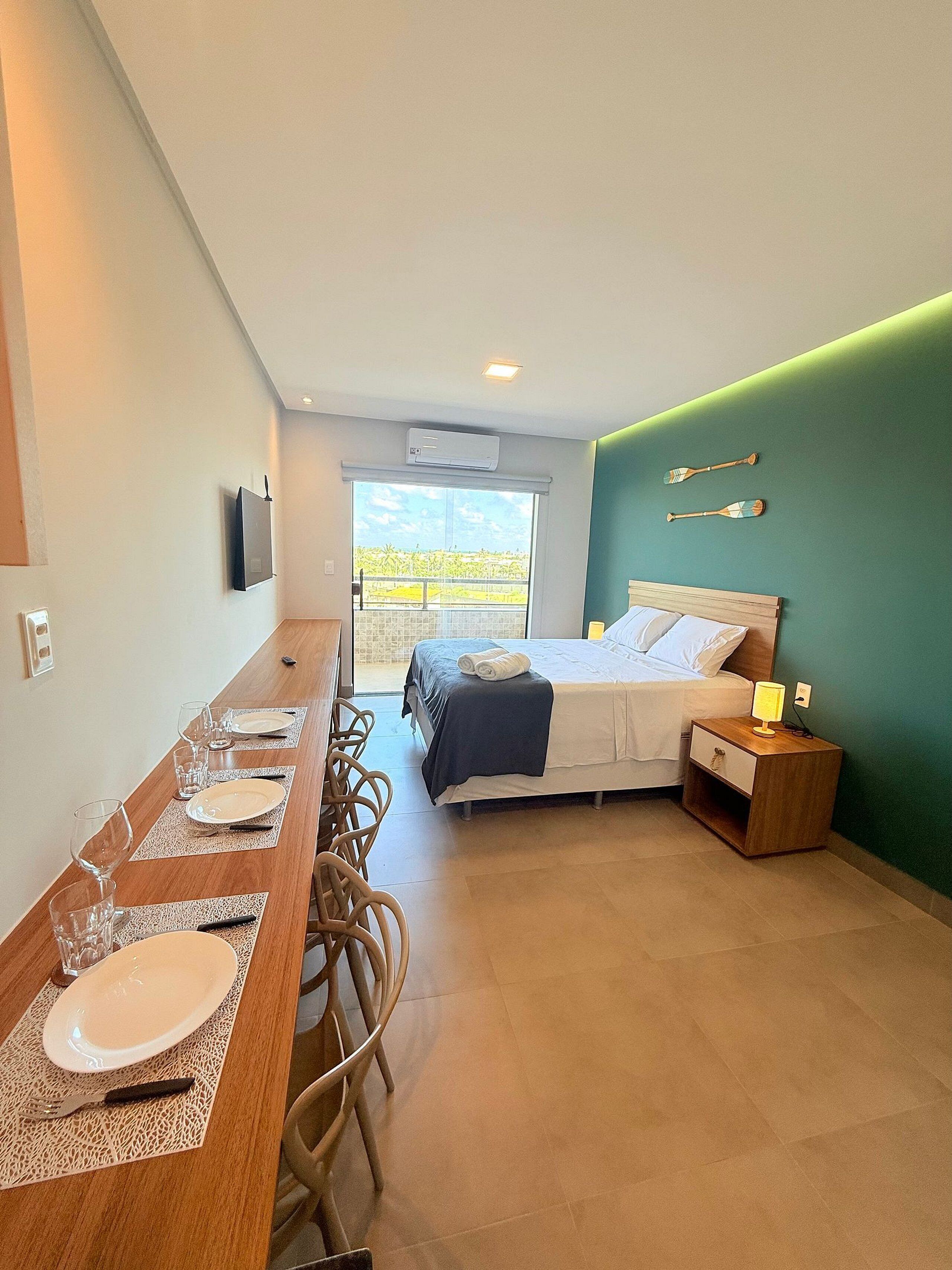 A room with a bed, a dining table set with plates and glasses, a TV mounted on the wall, and a view of greenery outside.