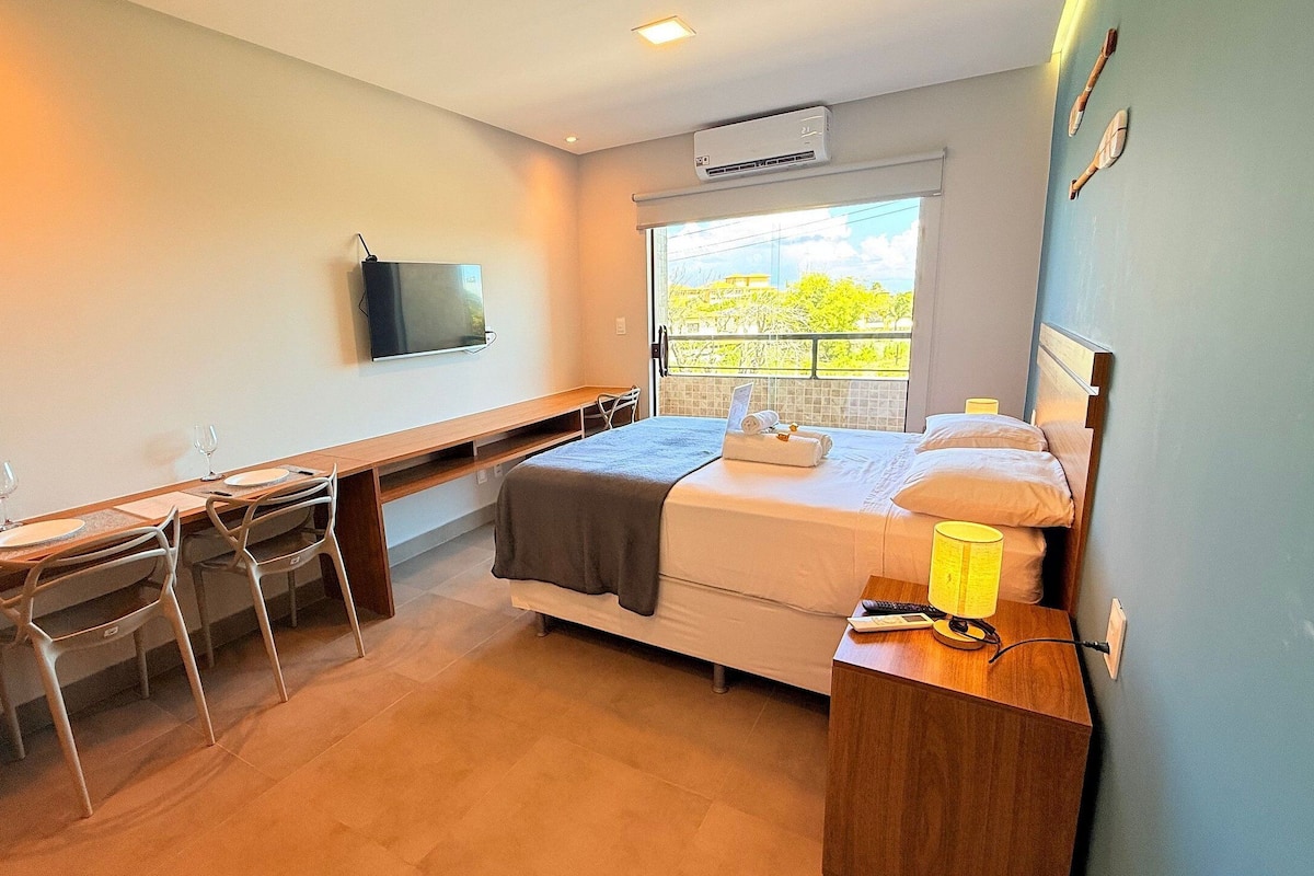 A compact hotel room with a bed, bedside table, desk, chairs, a TV, and a window with a view.