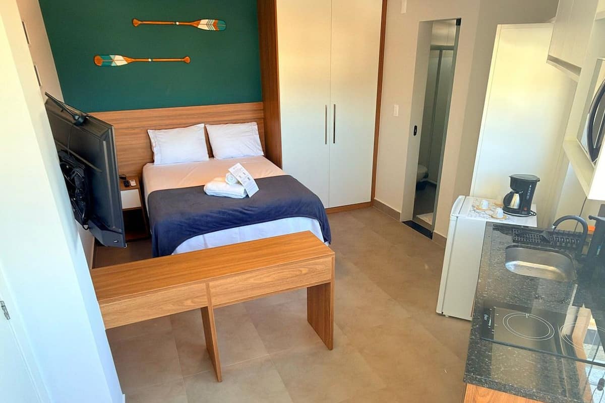 A compact hotel room with a bed, a small desk, a chair, a kitchenette, and a wardrobe.