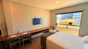 A hotel room with a bed, a desk with chairs, a TV on the wall, and a window with a view of buildings. - Stella Studio (Salvador)