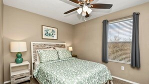 Un dormitorio con cama, mesita de noche, ventilador de techo, ventana con persianas e imagen colgada en la pared.