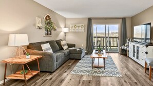 Una sala de estar moderna con un sofá modular, una mesa de centro de madera, un televisor y un balcón con mobiliario exterior.