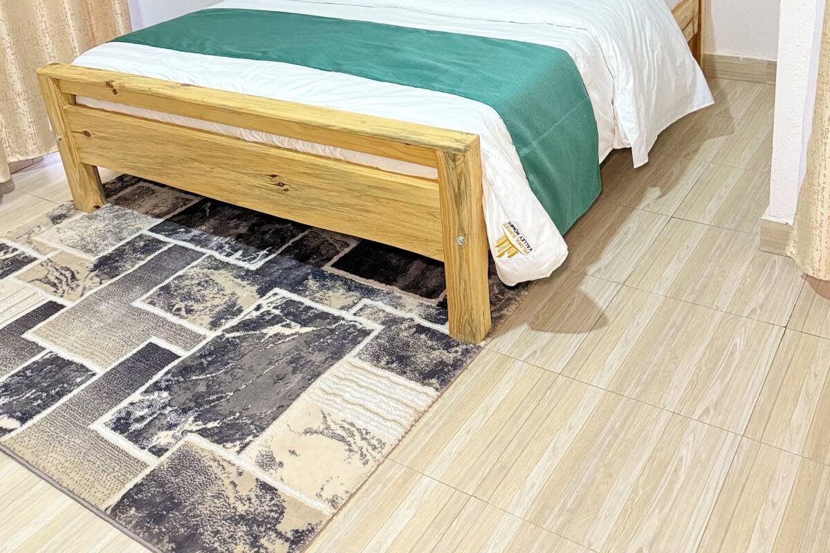 A wooden bed with a green and white bedspread, a patterned rug, and a plain wall.