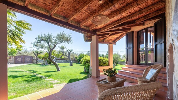 Un patio techado con mobiliario de mimbre, vista a un césped cuidado y un muro de piedra al fondo.